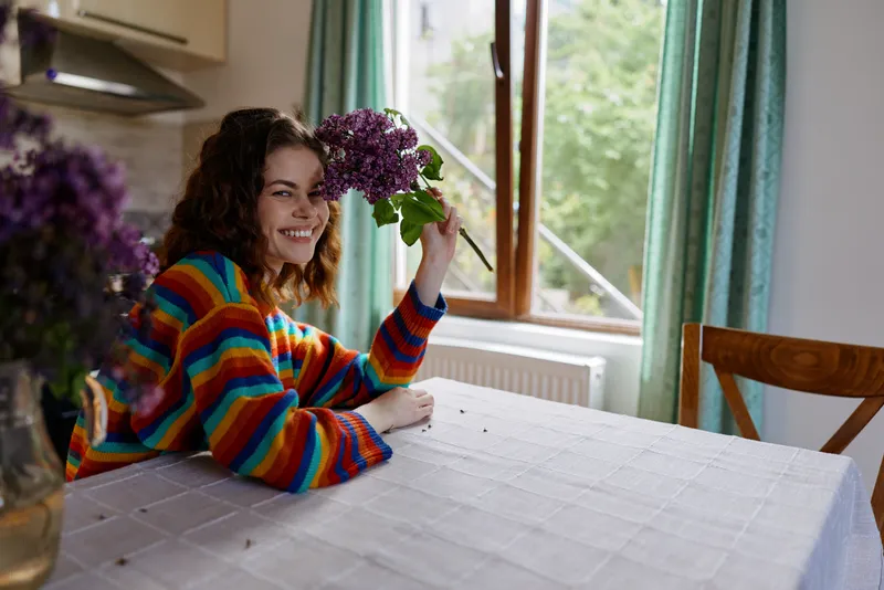 A young woman with curly hair sits at a bright kitchen table, smiling as she holds a bouquet of purple flowers, creating a warm, inviting moment of everyday happiness.