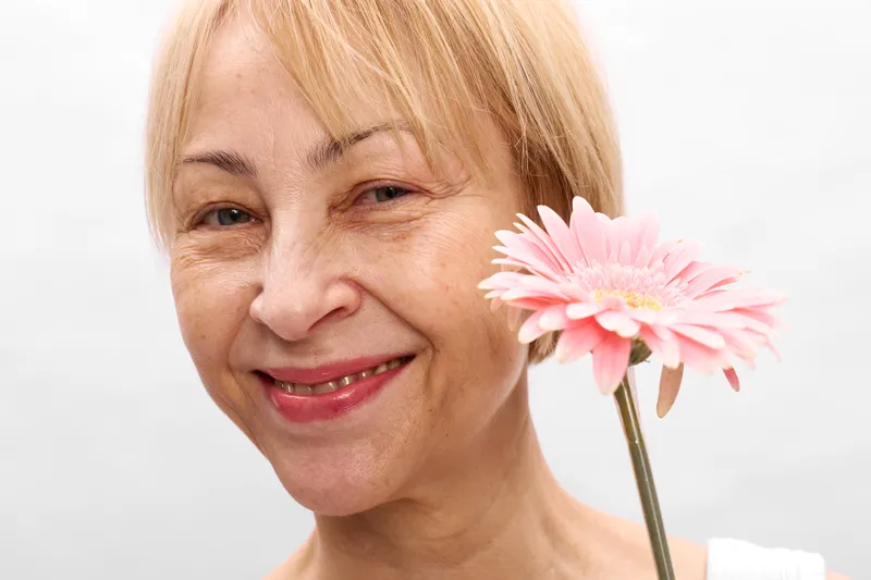 woman elderly smile flower pink gerbera closeup portrait, blonde headshot of a happy senior holding a single pink gerbera, white studio background, soft daylight, positive mood