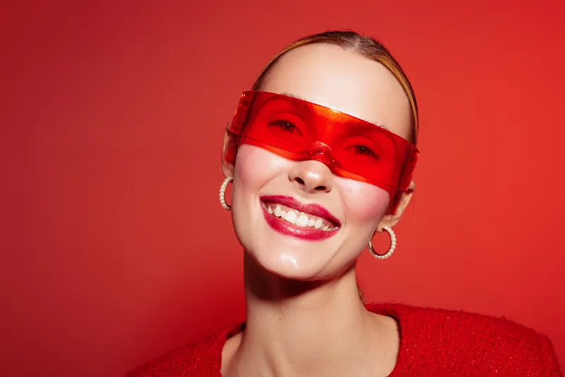 Smiling woman with red sunglasses and hoop earrings poses against red background. She wears a red top and has a bright, confident expression. Concept of fashion, style, and happiness.