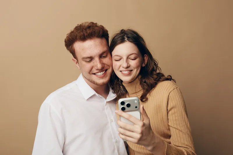 Smiling young couple takes a selfie together on smartphone. They stand close with happy expressions against a beige background. Concept of love, relationship, and modern technology.