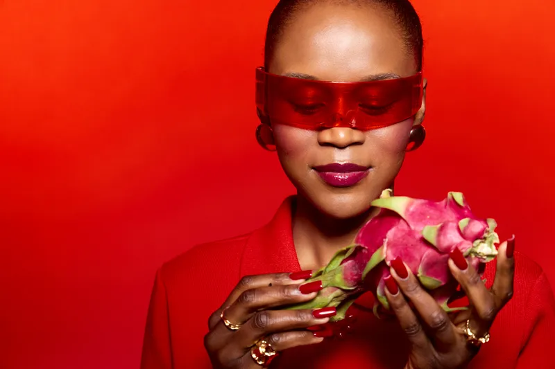 Dark skinned African woman wearing red sunglasses and red outfit holds a dragon fruit with manicured nails. Studio portrait on red background. Concept of fashion, beauty, and exotic fruit.