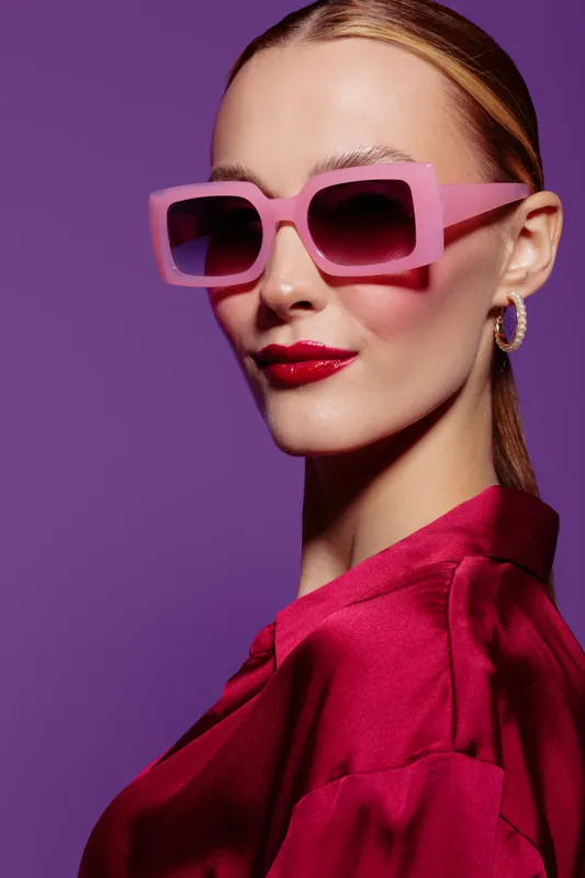 Stylish woman with pink sunglasses and red lipstick poses against purple background. Fashion portrait with glossy makeup and elegant red satin blouse. Concept of glamour and confidence.