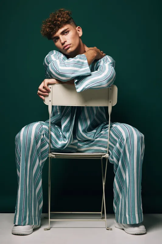 Young man with curly hair sitting on chair facing backward wearing striped pajamas looks thoughtful in a casual indoor relaxed pose with modern hairstyle and flat shoes.