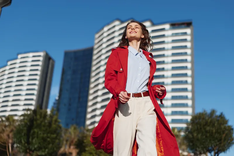 Young woman in a red coat walks confidently outdoors in an urban setting with modern buildings behind her, enjoying a bright sunny day with a joyful expression.