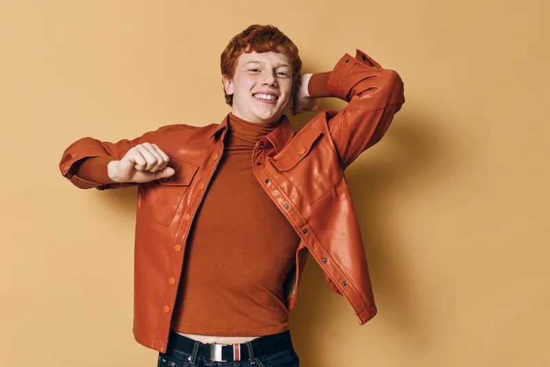 Young man with red hair smiling and dancing in orange jacket and sweater against beige background with casual style and joyful expression.