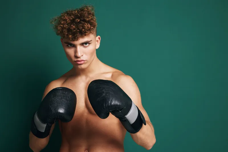 Young male boxer with curly hair wearing black boxing gloves in fighting stance against green background. Athletic shirtless man training for boxing or martial arts workout.
