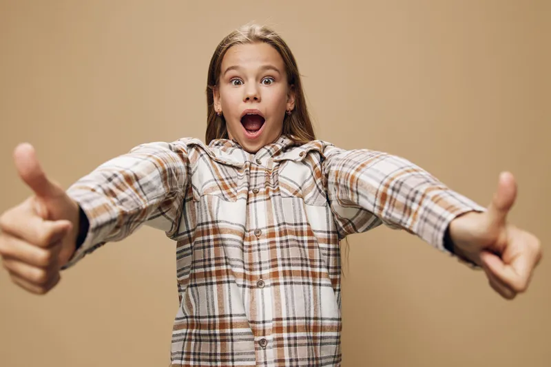 Girl wearing plaid shirt showing thumbs up with excitement and surprise on face, posing on beige background with open mouth and wide eyes, casual style and positive emotion.