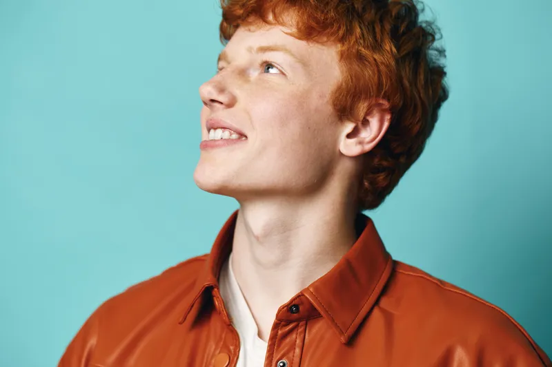 Young man with red hair and light skin wearing a brown leather jacket looking upwards smiling on blue background. Casual portrait with natural expression and hairstyle.