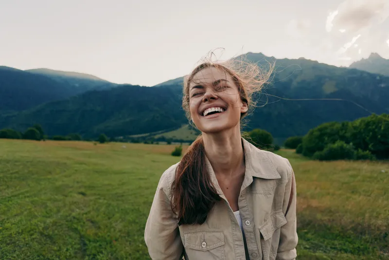 Happy woman with long hair smiling outdoors in a green field with mountains in the background. Natural light highlights a carefree lifestyle and joyful mood.