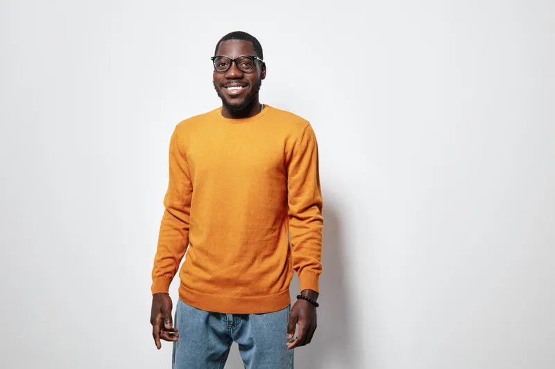 African American man with dark skin wearing orange sweater and glasses smiling standing against white background in casual outfit and relaxed pose.