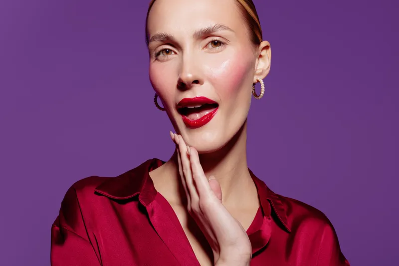 Close-up of a fashionable woman with red lipstick and hoop earrings posing against purple background. Concept of beauty, makeup, style, and confident expression.