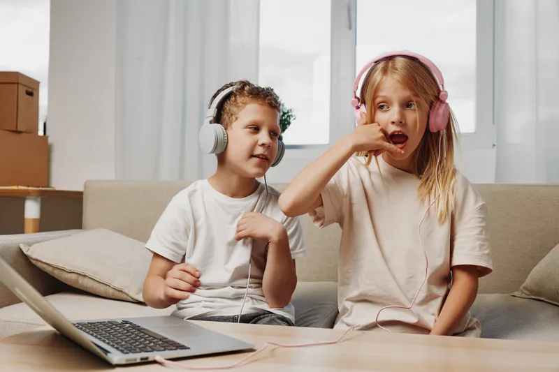 Two children with headphones sit on a sofa using a laptop, enjoying music and interacting in a bright living room. Concept of childhood, technology, and entertainment.