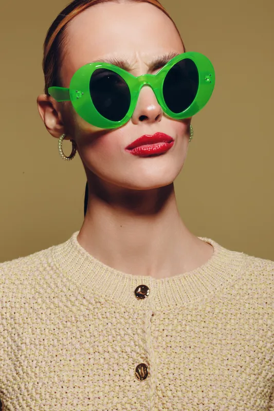 Stylish woman with green oversized sunglasses and red lipstick wears beige knitted sweater. Close-up portrait on neutral background. Concept of fashion, accessories, and confident attitude.