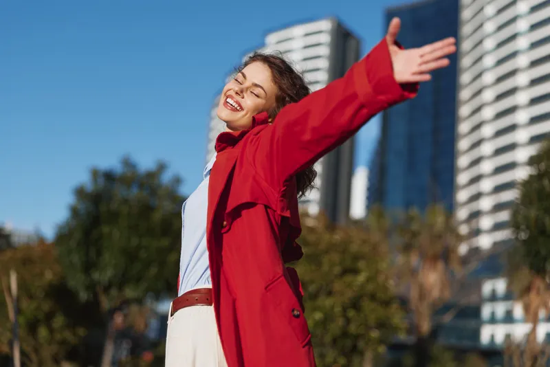 Happy woman outdoors wearing red coat, arms wide open, eyes closed, smiling joyfully, urban background, sunny day, casual outfit, carefree mood