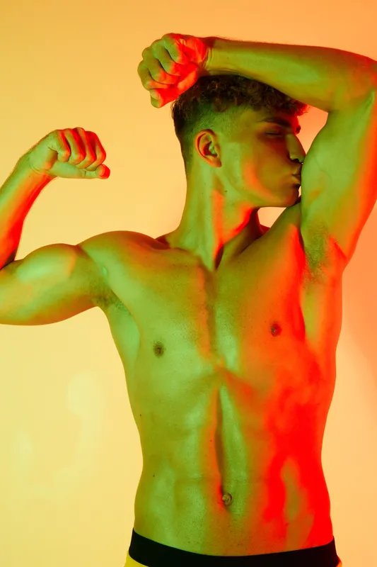Muscular man posing shirtless with flexed biceps and toned body under colorful lighting for fitness, strength, health and athletic concept in studio portrait.