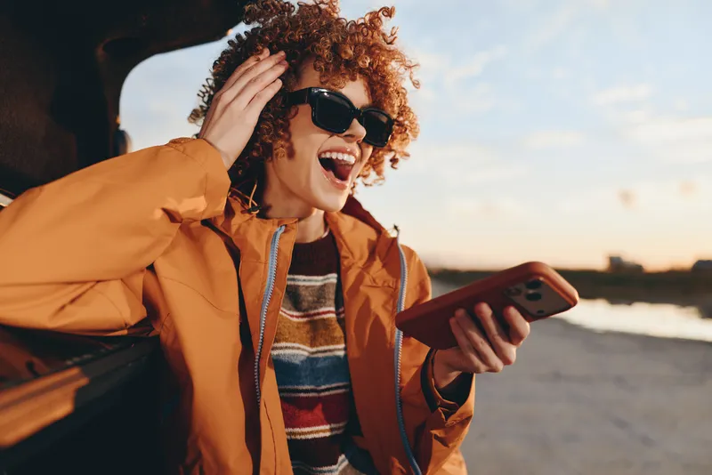 Woman lifestyle, curly woman outdoors, laughing in sunglasses while holding smartphone, warm sunset light on face, casual jacket and striped top, seaside promenade background, candid cheerful moment