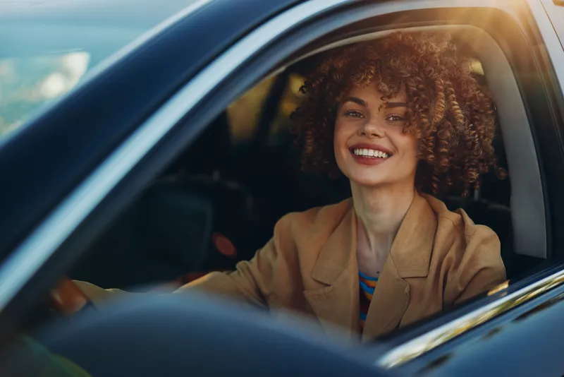 Young woman drives a car, smiling through the open window, retro film color tones, casual blazer, carefree mood, candid lifestyle portrait in warm sunlight