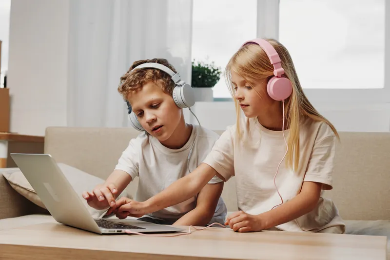 Two children wearing headphones use laptop together at home, focused on screen. Concept of learning, technology, childhood and cooperation in casual setting.