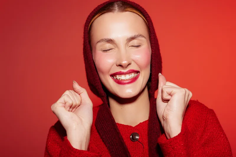 Happy young woman with closed eyes and bright red lipstick smiles joyfully wearing a red hooded sweater. Concept of happiness, beauty, fashion, and positive emotions on red background.