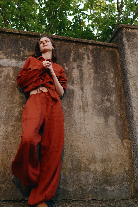 A woman in a red jumpsuit leans against a weathered concrete wall outdoors, looking upward with a calm, thoughtful expression. Concept for urban style and resilience