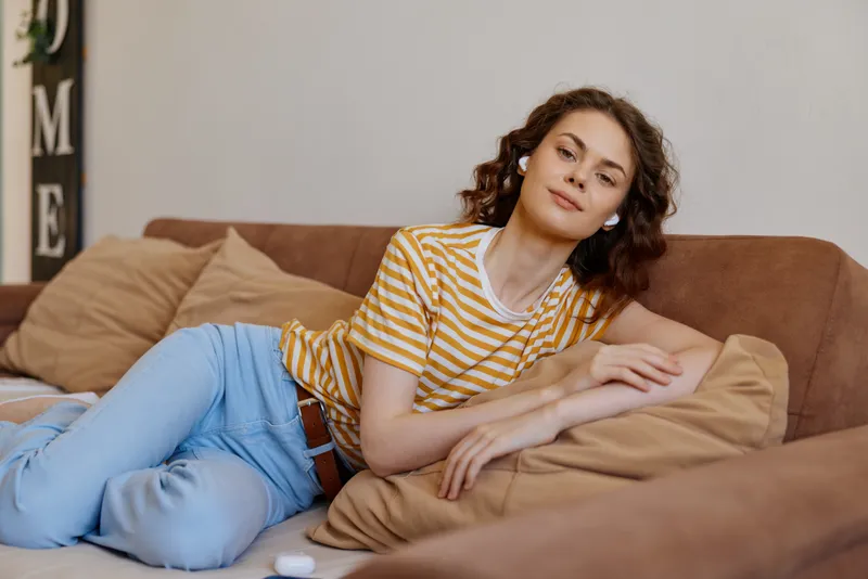 Relaxed woman lounging on a beige sofa wearing a striped yellow shirt and light blue jeans in a cozy home setting. The scene conveys casual comfort, selfcare, and everyday wellness.