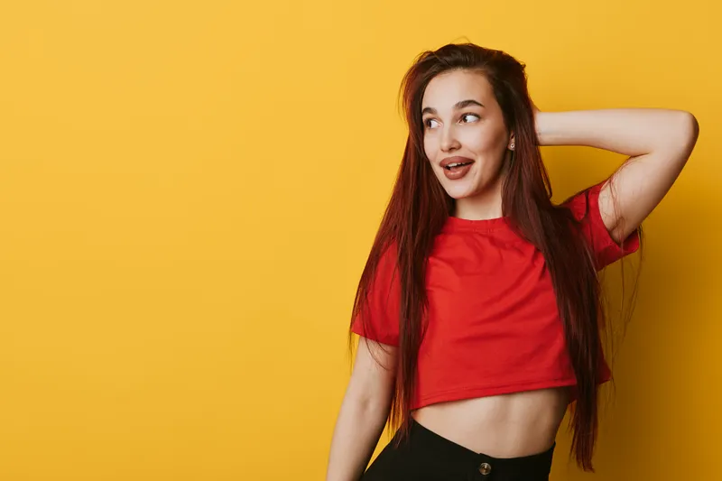 Young woman with long hair wearing red crop top posing with hand behind head looking to side on yellow background. Casual style, beauty and confident expression in studio.