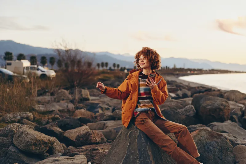 Woman lifestyle portrait at sunset on rocky beach, curly haired woman in orange jacket, striped shirt walks among rocks, distant mountains on horizon, warm outdoor light