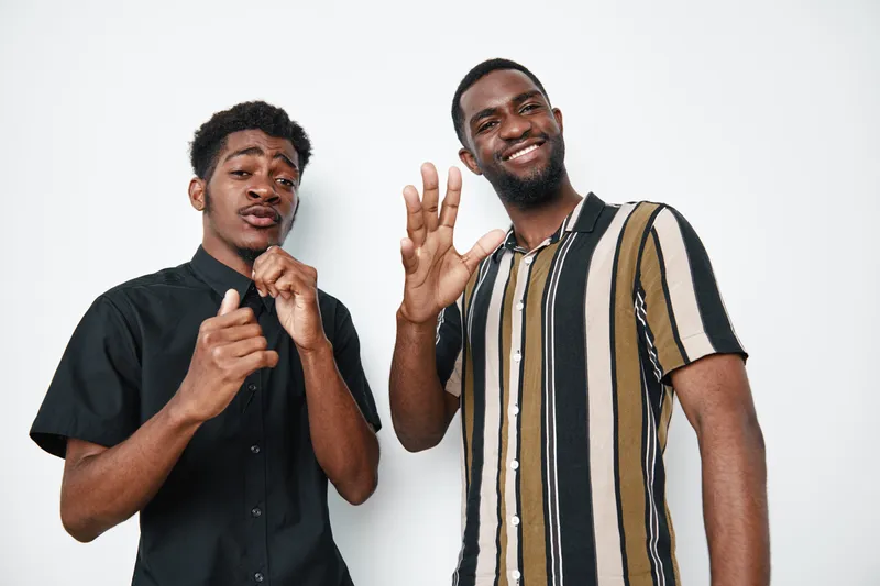Two young black men with dark skin posing and smiling on white background. Casual clothing, friendship and positive emotions with gesture and confident expressions in studio.