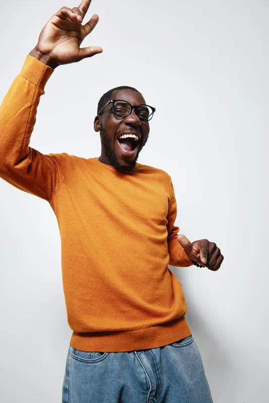 African American man with dark skin wearing glasses and orange sweater smiling and dancing with raised hand in casual jeans against white background in studio. Joyful, happy, energetic.