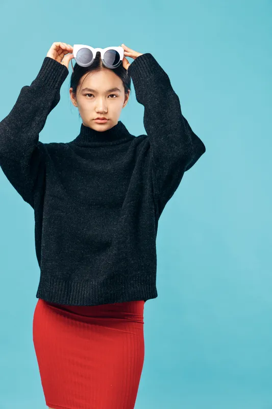 Young woman in black sweater and red skirt adjusting round sunglasses on head against blue background. Fashion, style, portrait and casual outfit with confident expression in studio.