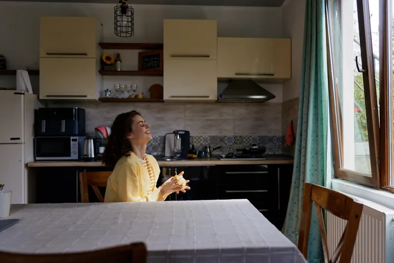 Young woman in a yellow cardigan sits at a kitchen table smiling while looking toward a sunlit window in a cozy home kitchen, creating a warm, inviting lifestyle moment.