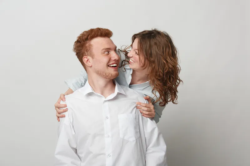 Happy young couple with red hair and curly hair smiling and hugging each other on a light background. Concept of love, friendship, relationship, and positive emotions.