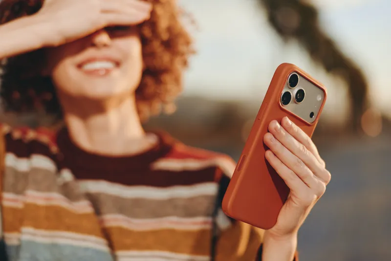 Woman lifestyle portrait, woman with curly hair showing a smile, holding smartphone outdoors with orange case, casual striped sweater, soft natural light, warm mood, communication, blurred background