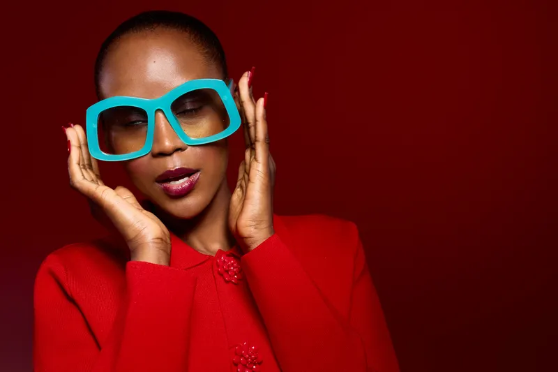 Portrait of a black woman with dark skin wearing large turquoise glasses and a red coat. She poses confidently against a red background, showcasing fashion and style concepts.