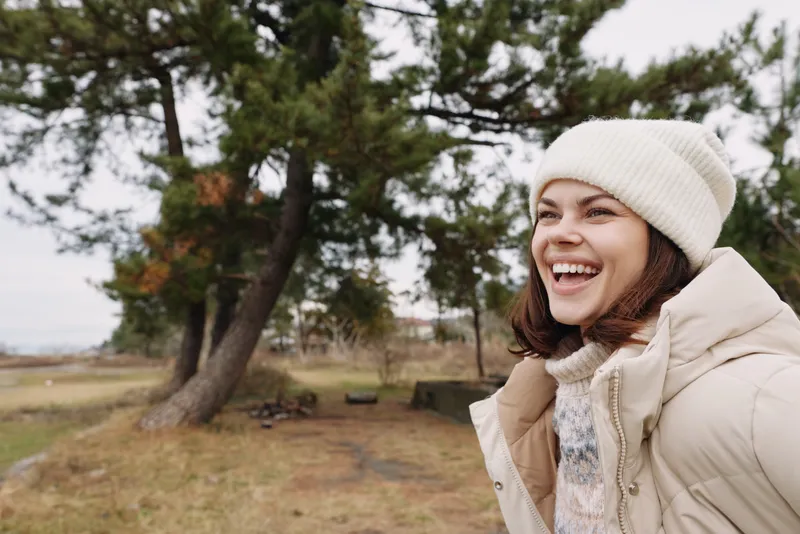 Outdoor woman, smiling portrait in a winter jacket and beanie, enjoying the cool breeze in an open park with trees, space and a joyful mood, natural light
