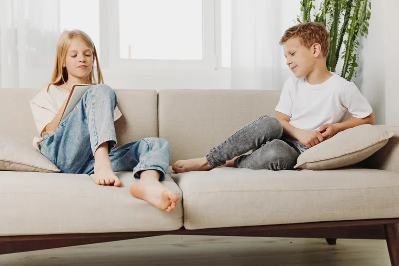 Two children relax on a modern beige sofa in a bright living room, barefoot and casually dressed. The boy holds a cushion while the girl looks down thoughtfully, calm atmosphere.