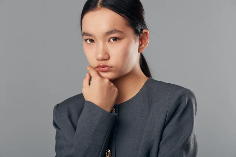Young Asian woman with serious expression wearing gray professional jacket posing thoughtful and confident with hand near face on solid gray background in studio portrait.
