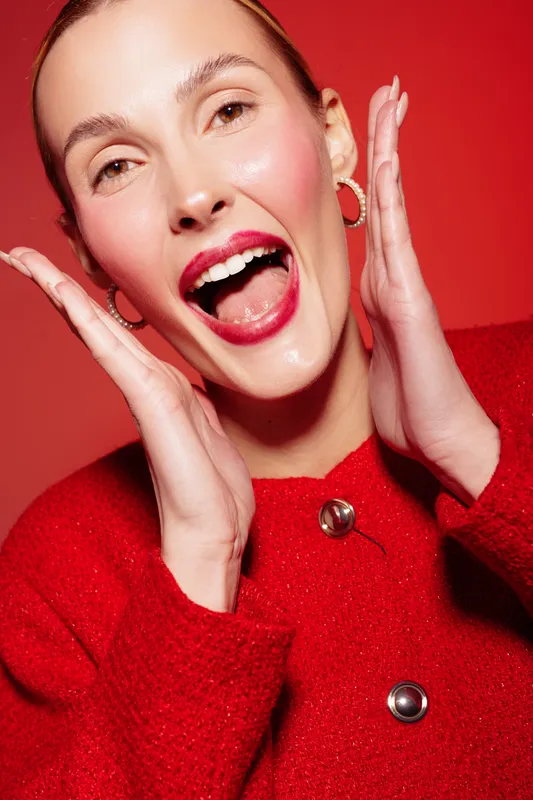 Happy woman with red lipstick and red sweater smiles widely with hands near face on red background. Concept of joy, fashion, beauty, and vibrant emotions in studio portrait.