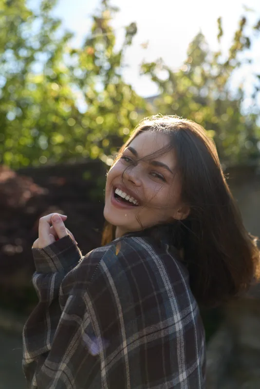Woman embraces outdoor lifestyle with a joyful smile, wearing a plaid shirt in warm sunlight, captured in a candid portrait against a natural backdrop