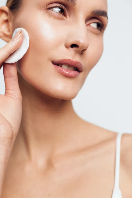 Young woman removes makeup with a cotton pad in a bright studio, close-up on glowing skin and a calm, clean mood.