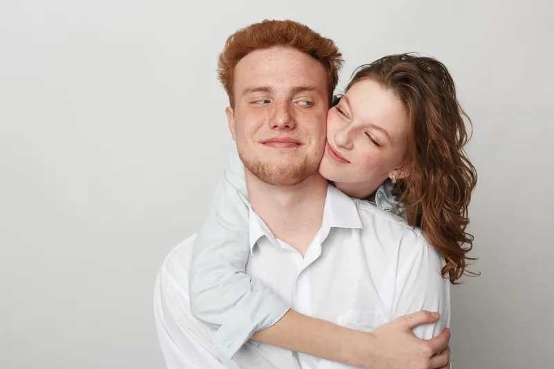 Young couple in casual clothes embraces and smiles gently against a plain background. The woman hugs the man from behind showing affection and warmth. Concept of love and relationship.