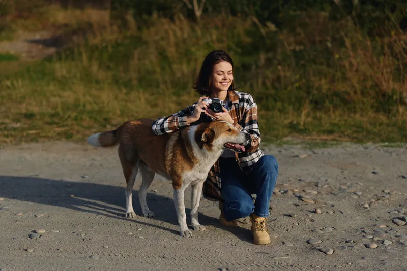 Woman crouching beside a friendly dog on a sandy path, camera in hand, capturing a candid outdoor moment amid grass and open nature, warm light