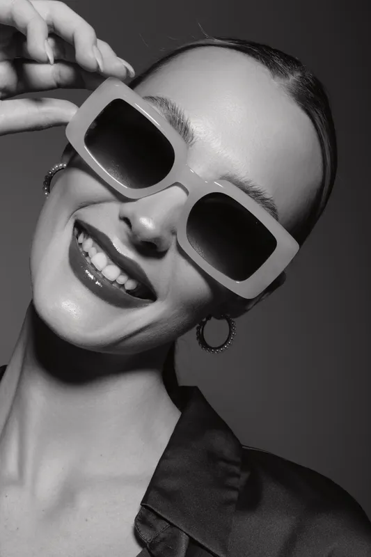Young white woman with pale skin wears large sunglasses and hoop earrings smiling in studio black and white portrait.