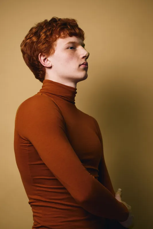Young man with red hair wearing brown turtleneck, posing in profile against beige background. Portrait of thoughtful expression, fashion style and casual clothing with side view.