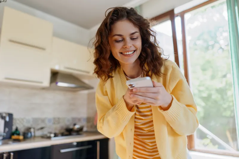 Smiling woman in a yellow cardigan using a smartphone at home in a bright kitchen. Casual lifestyle, warm mood, everyday technology, relaxed morning routine, candid indoor moment, daylight glow.
