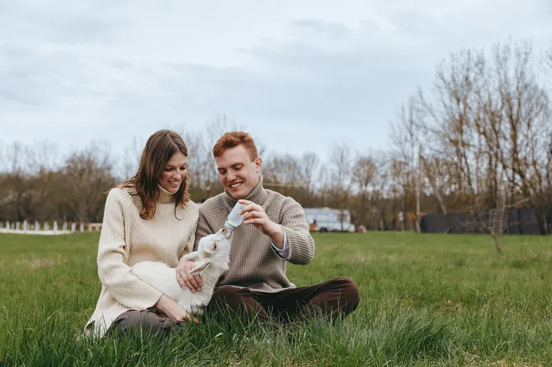 Happy couple enjoys a gentle moment outdoors with a playful goat kid, sitting on the grass in a park, sharing smiles, cozy sweaters, natural light, rustic lifestyle and warm connection.