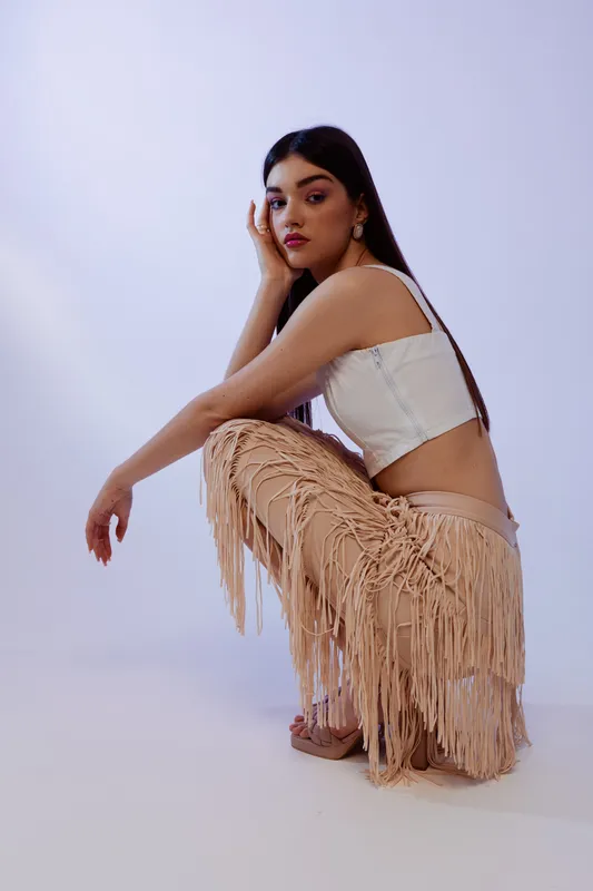 Young woman in a white crop top and beige fringed pants poses in a studio. She looks confidently at the camera with a calm expression. Concept for woman beauty fashion.