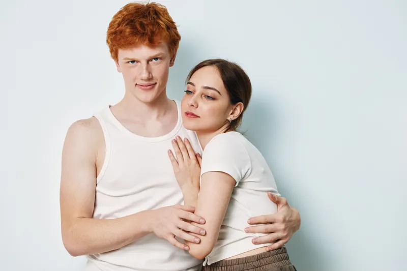 Couple, young man and woman hugging closely with emotional expression in casual white clothing on light background in studio. Relationship and affection.