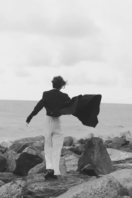 Man standing on jagged rocks by the sea, coat billowing in wind, solitary figure facing the horizon in a monochrome scene, dramatic and resilient