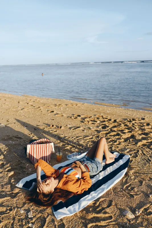 Young woman in denim shorts and orange jacket relaxes on a striped towel on the sandy beach near calm water under clear sky during sunny day.
